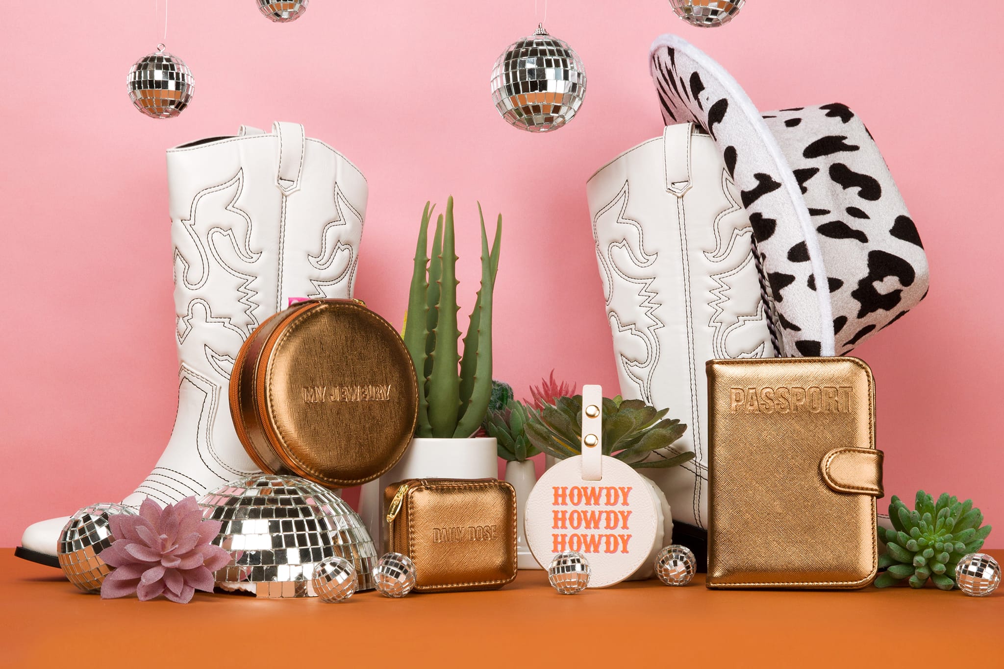Go West collection accessories are displayed: brown metallic-colored round jewelry case, daily dose rectangle pill case, and passport case, along with "howdy" luggage tag. Accessories are surrounded by cacti, cowboy boots, and disco balls.