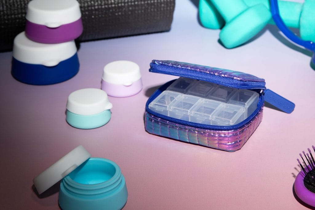 A shiny, metallic purple pill case is open showing its days of the week compartments. It sits next to silicone stacking jars that are blue and purple.